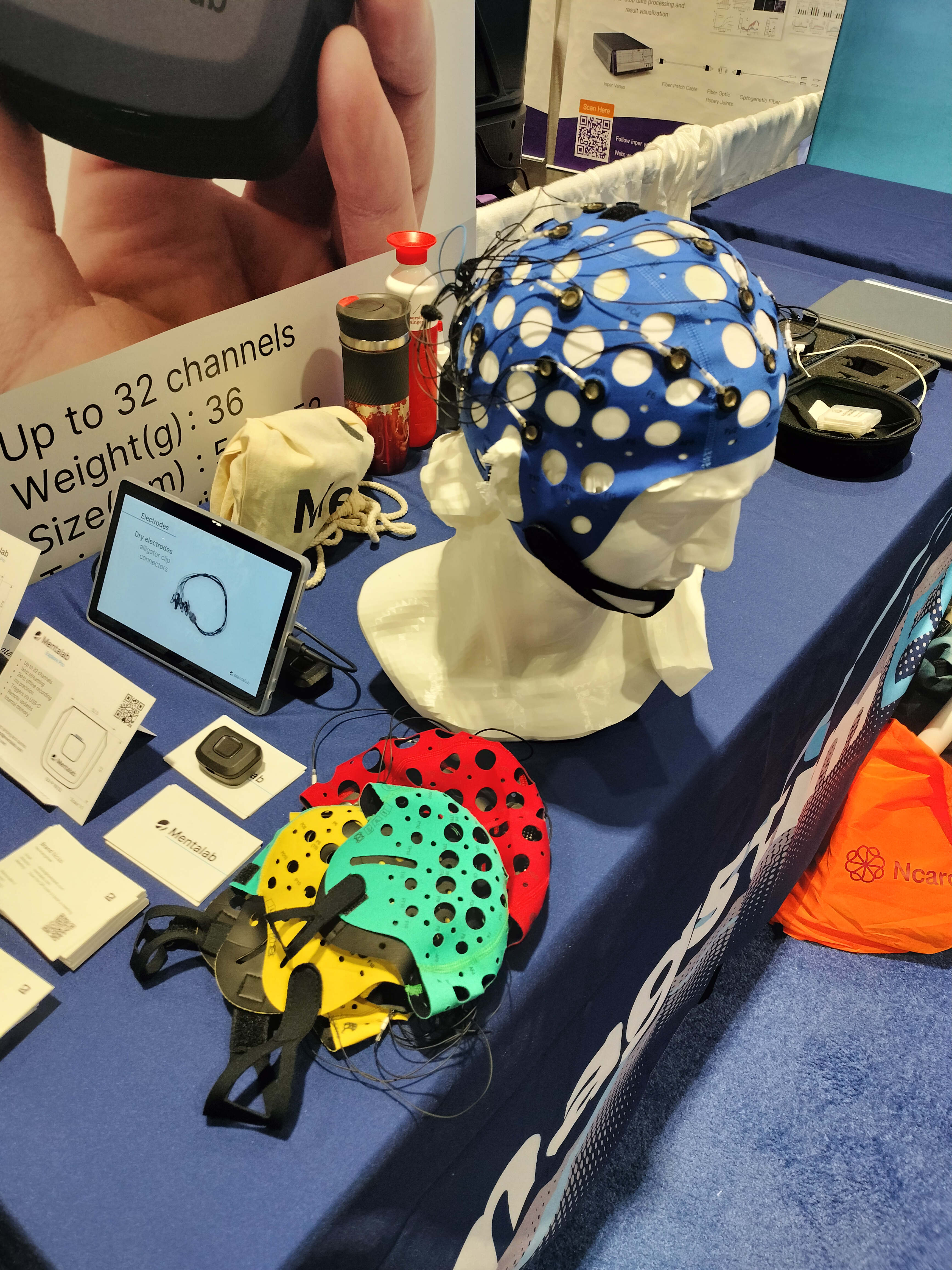 On a table, a mannequin head displays a blue EEG cap that covers the entire head except for the face. The cap is perforated with a pattern of holes, resembling swiss cheese, with electrodes placed in the gaps. Wires run from the electrodes to the back of the cap. Lying flat on the table next to the mannequin are additional EEG caps in red, yellow, and green, along with a tablet and a small, box-like electronic device for data acquisition.