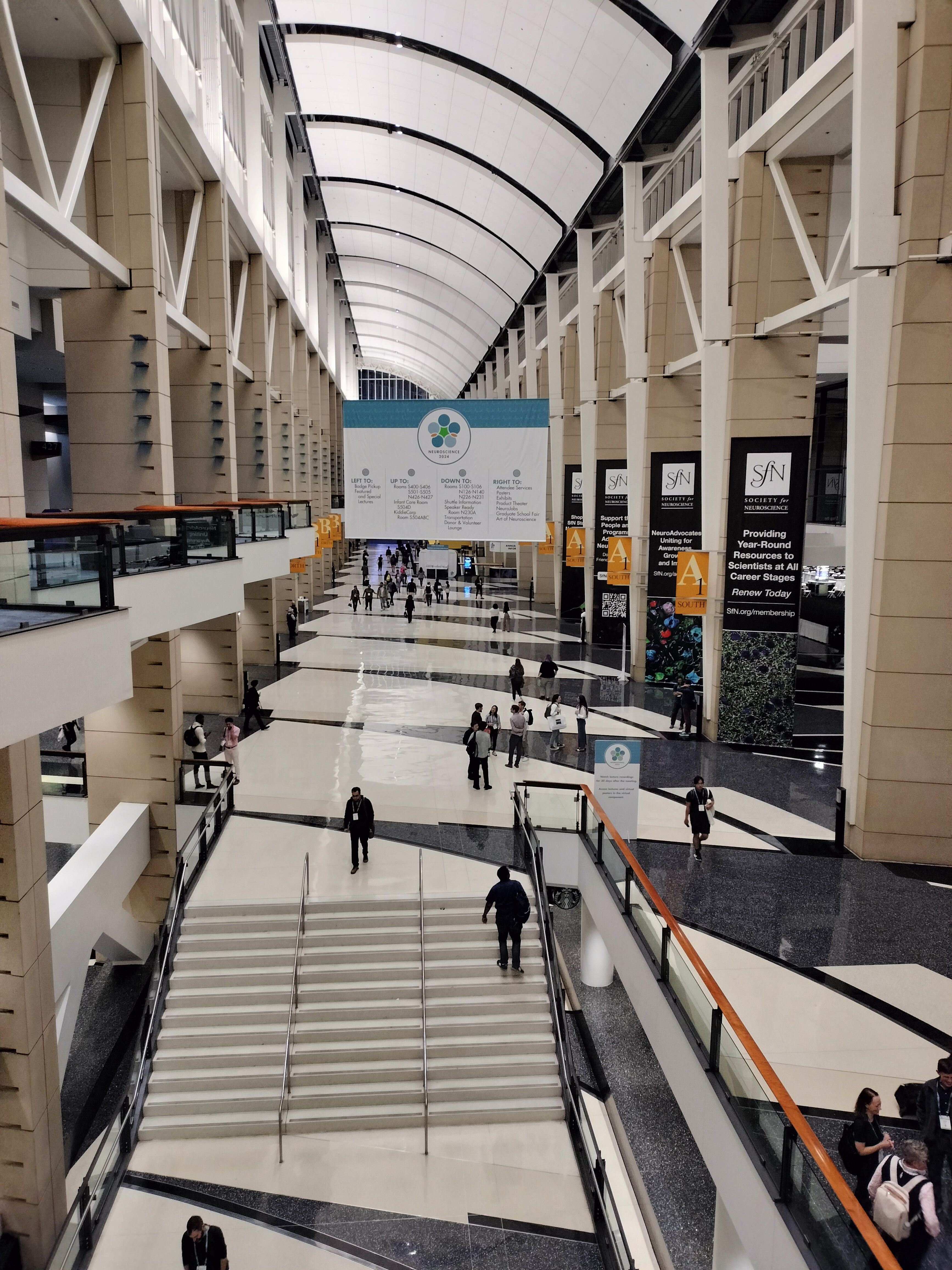 A wide, expansive hallway with a very high, architecturally distinct ceiling forms the Grand Concourse. Large pillars line both sides of the walkway, which directs people to the North building on the right and the South building on the left. High above, a large banner for the SfN conference hangs from the ceiling. The ceiling itself is a segmented drop-down design, featuring large, concave tiles. On the left side, balconies overlook the bustling concourse below.