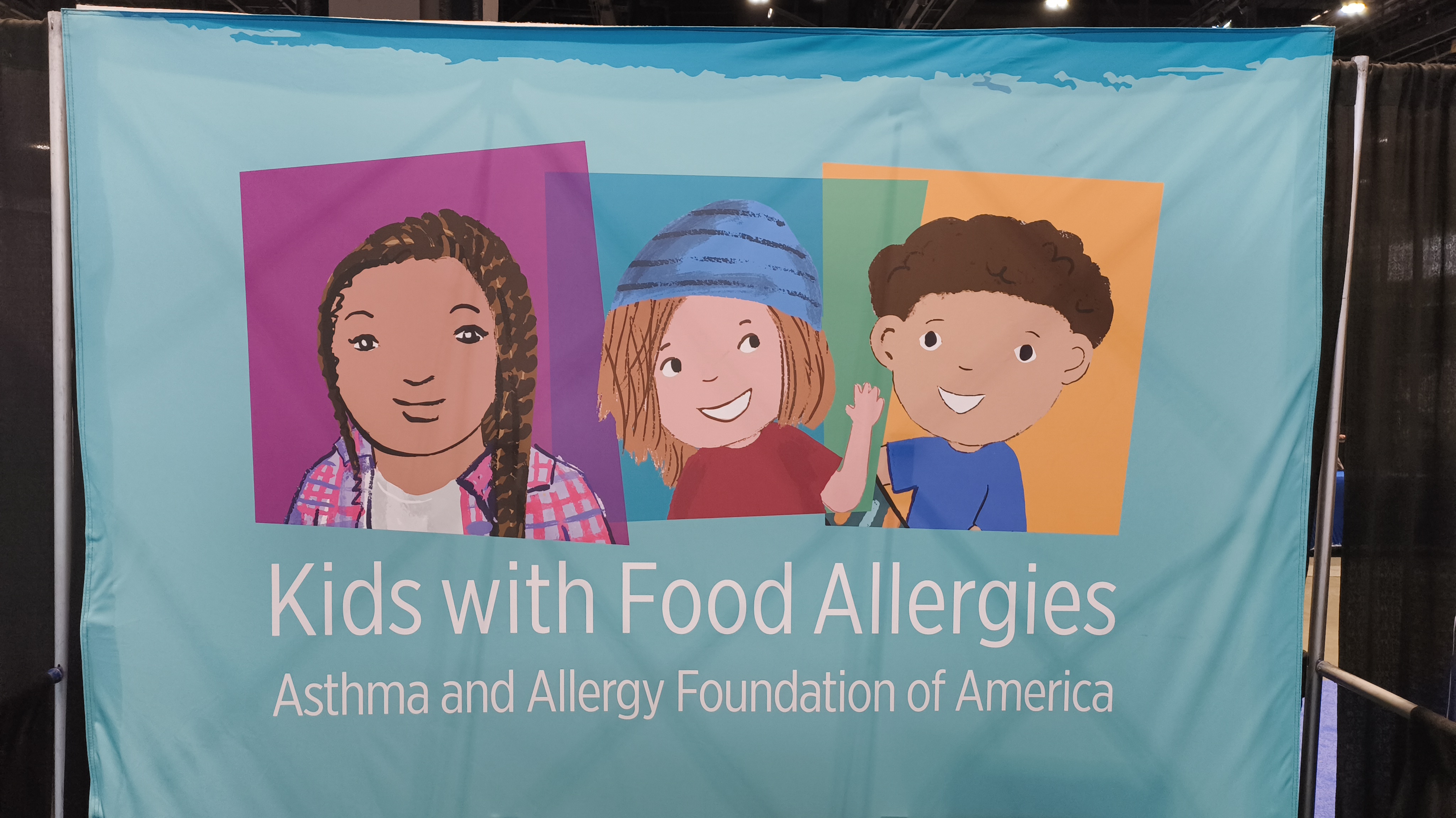 A backdrop of the exhibitor booth for the Asthma and Allergy Foundation of America (A.A.F.A.), an N.G.O. nonprofit. Printed in white on the front is the title “Asthma and Allergy Foundation of America.” To the left of the title is the AAFA logo: the lowercase letters “aafa” surrounded by many white dots. On the right side of the table, many pamphlets are displayed face up, partially overlapping. In the center, a stand props up more brochures. On the left side is a sign-up sheet. Behind the table sit two chairs in front of a light blue backdrop. The backdrop features three cartoon portraits of children side-by-side. The leftmost portrait features a girl with tan skin and black hair, wearing a pink open shirt over a white blouse against a purple background. The center portrait features a fair-skinned girl with brown hair and a blue beanie, wearing a red shirt against a blue background. Where the two girls’ backgrounds overlap, the color is dark purple. The rightmost portrait features a fair-skinned boy with black hair, wearing a blue shirt against a yellow background. The overlap between the center girl’s and the boy’s backgrounds is green.